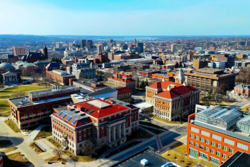 Aerial view of a university campus in an urban environment, with classic academic buildings in the foreground and a modern cityscape in the background.