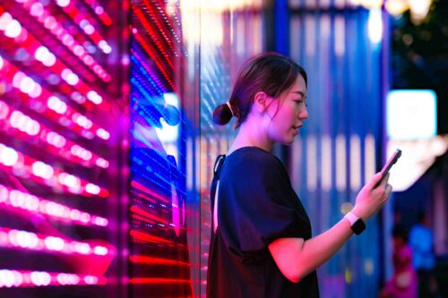 A woman with a low bun stands holding a smartphone, illuminated by pink and blue lights in a vibrant urban setting.
