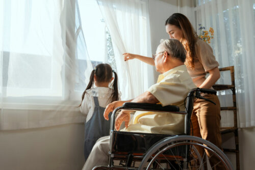 An adult woman gestures toward a window where she stands next to an older man in a wheelchair and a young girl with pigtails.