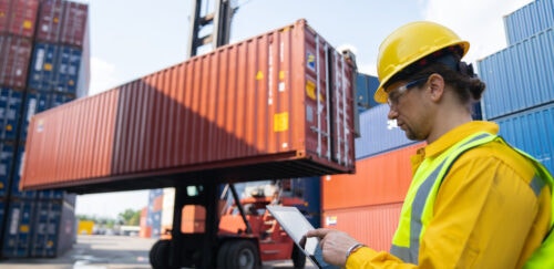 Worker in safety gear uses a tablet while a container handler lifts a red shipping container in a shipping yard.