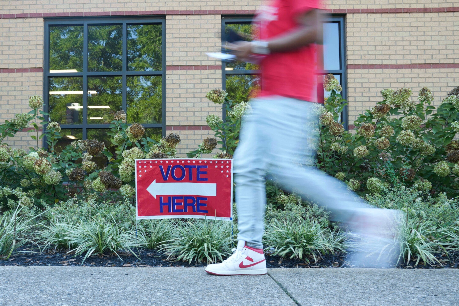 A red sign reading VOTE HERE with an arrow is placed in the landscaping between a sidewalk and a building. A man in a red shirt, light-colored pants, and white sneakers walks in the direction of the arrow, blurred by motion.
