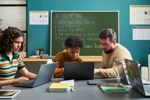 Caucasian man assisting Black teenager and Caucasian teenage girl working on laptops in classroom setting, chalkboard with AI prompting instructions visible in background.
